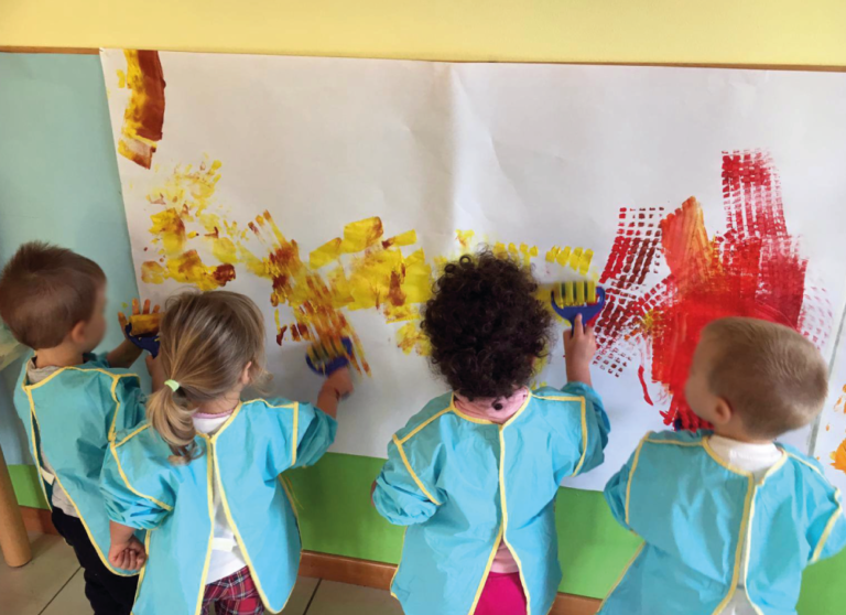 Bambini che dipingono con le tempere un cartellone nel salone dell'Asilo Valli.
